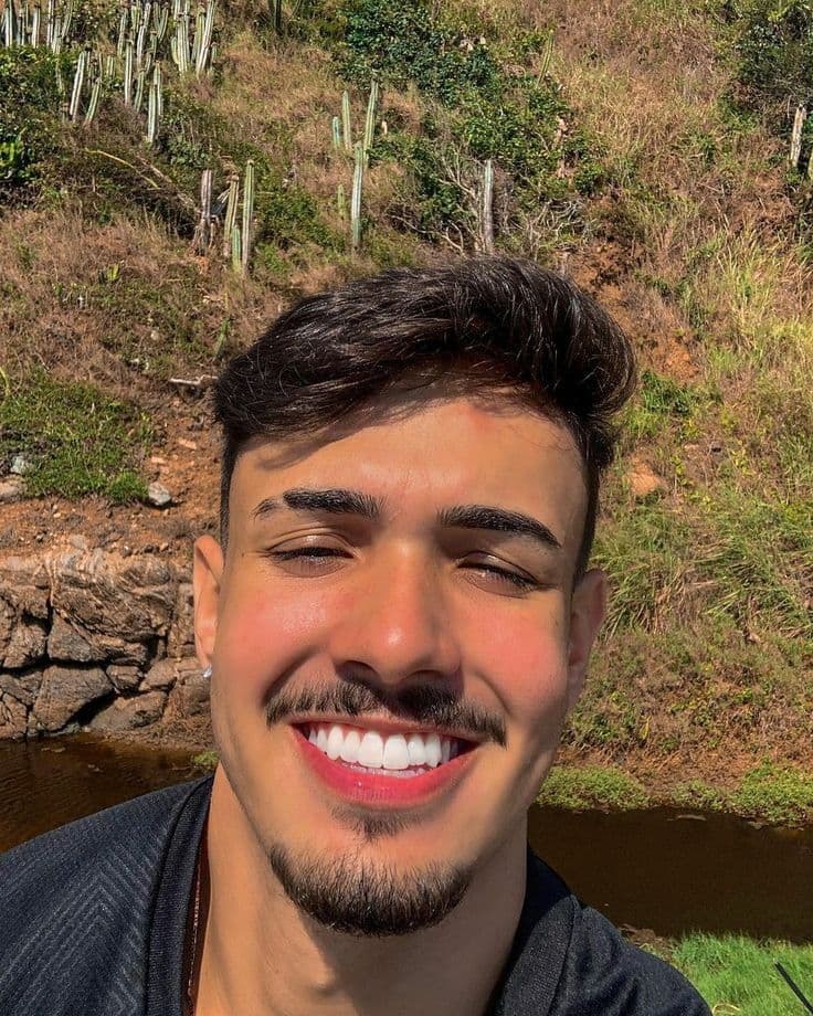 Homem jovem sorridente com bigode e cavanhaque em frente a uma colina ensolarada com cactos.