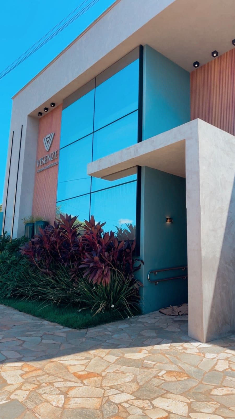 Fachada da Visenze Medicina Integrada com janelas espelhadas, detalhes em madeira e plantas vermelhas.