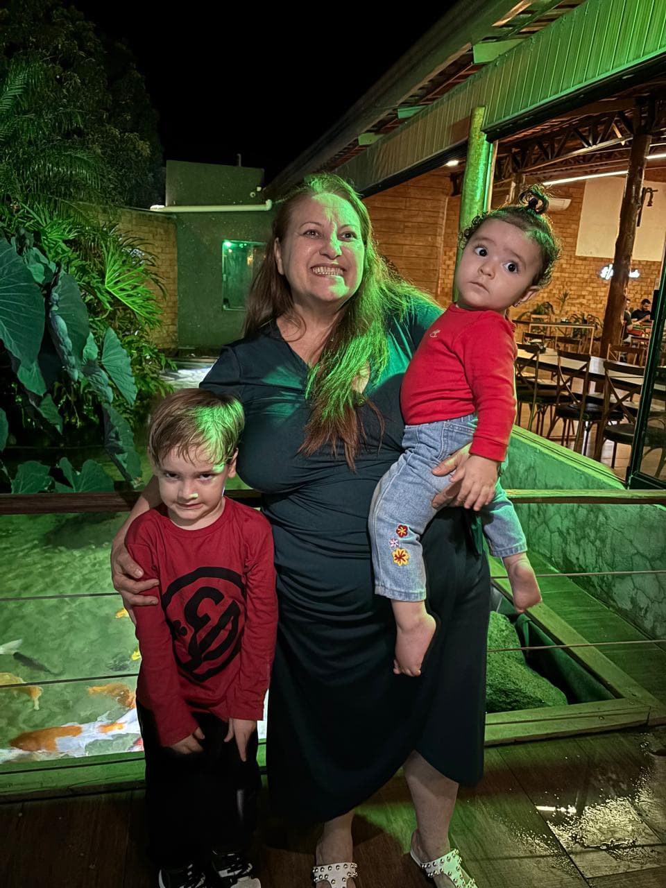 Mulher sorridente segura bebê e menino ao lado de lago com carpas coloridas à noite.