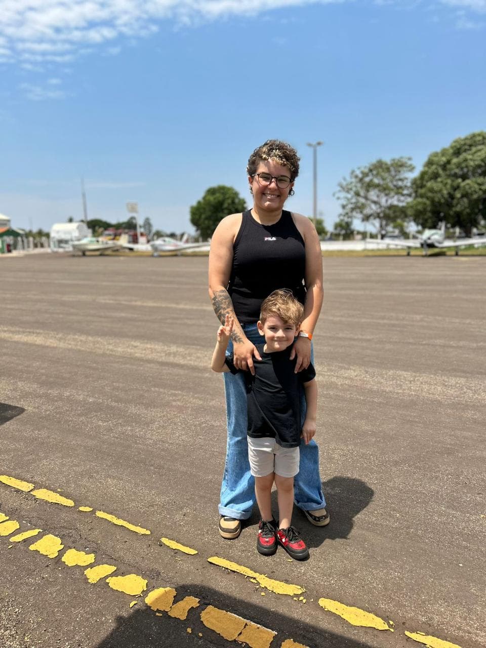 Uma mulher e um menino sorrindo em uma pista de pouso com aviões ao fundo.