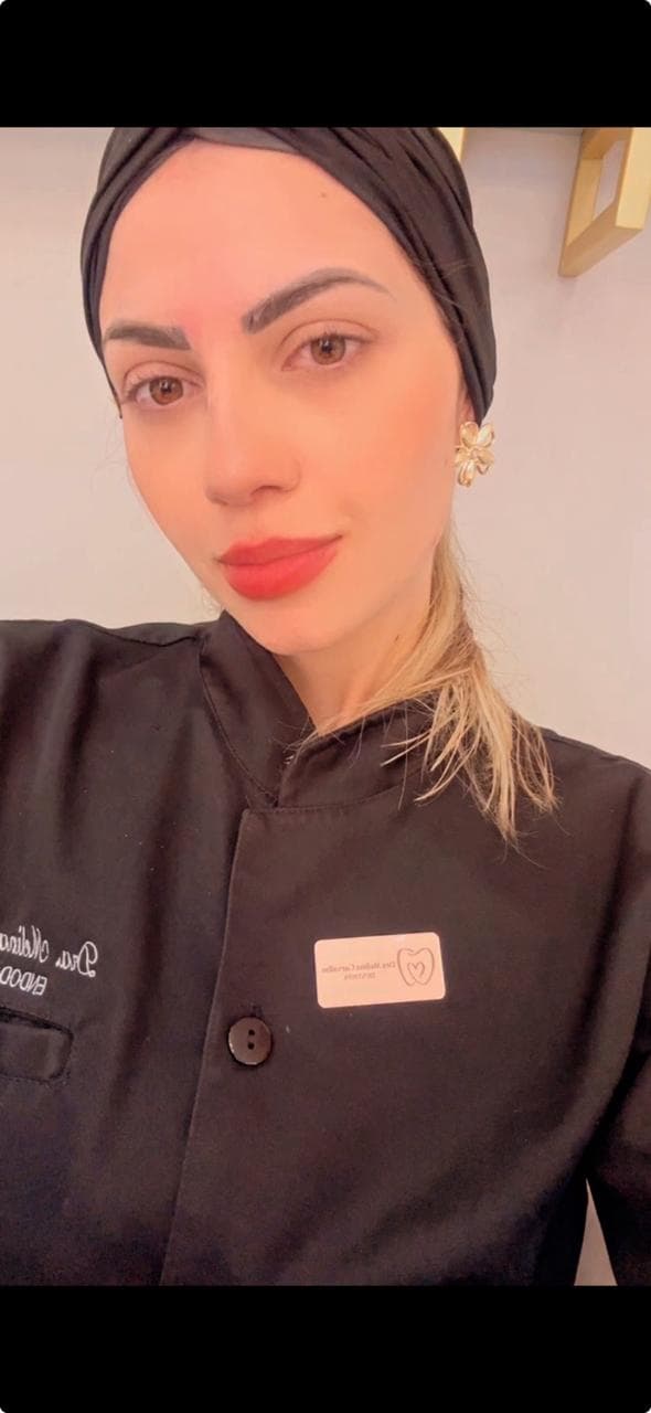 Mulher com uniforme odontológico preto, turbante, batom vermelho e brincos de flor dourados.