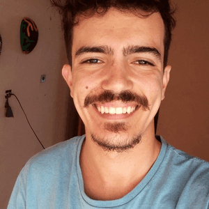 Homem jovem sorridente com bigode e cavanhaque, vestindo camiseta azul clara em ambiente interno.